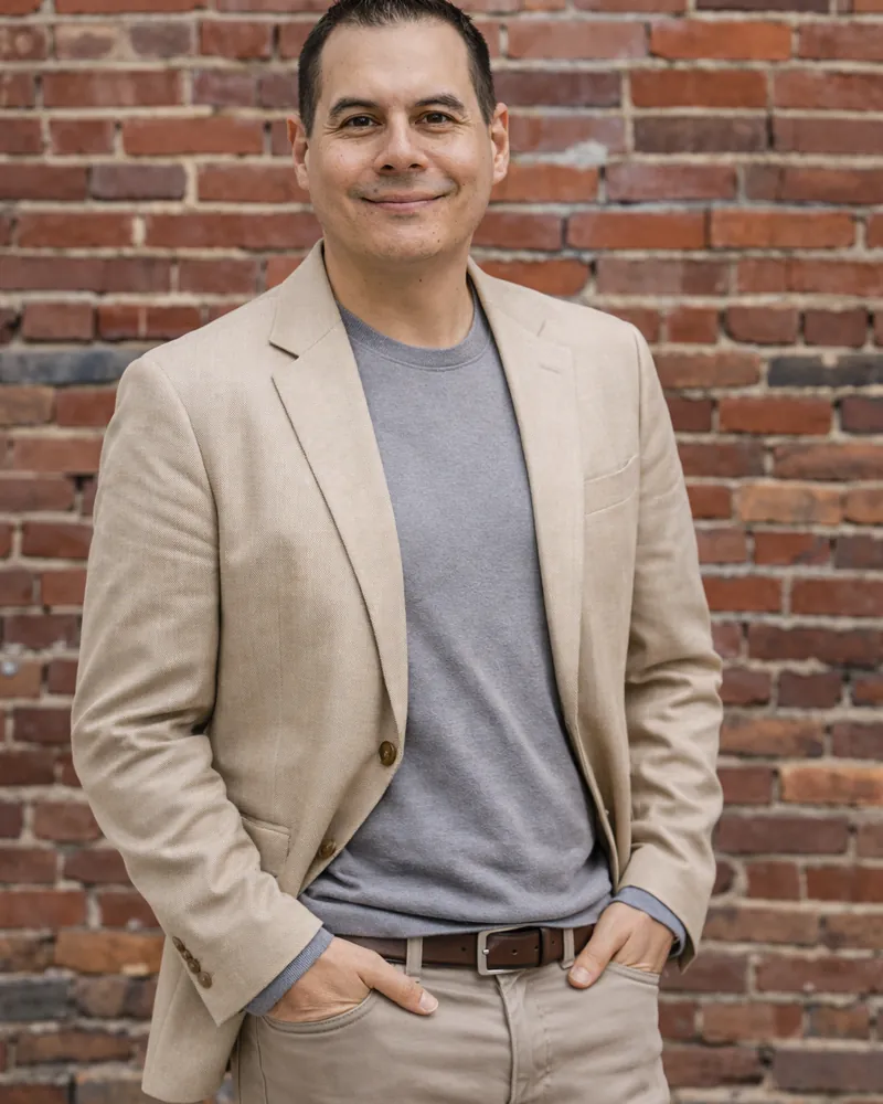 Nate Crandell standing in front of a red brick wall, wearing a tan blazer